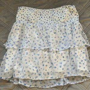 Vit blommig volangkjol Vero Moda - Supersöt kort kjol från Vero Moda i vitt med blått blommönster. Kjolen har flera volanger och smock i midjan för en bekväm passform. Perfekt för sommarens alla tillfällen och ger en fräsch och romantisk vibe. Den är i L men passar folk som har S-XL ändå för den anpassar sig. 