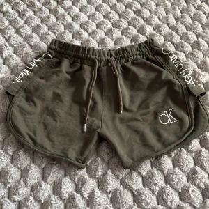 Olivgröna shorts Calvin Klein  - Snygga olivgröna shorts från Calvin Klein med vit logga på ena benet och Calvin Klein-tryck längs sidorna. De har elastisk midja med snörning och är tillverkade i mjuk bomull. Perfekta för chill dagar eller träning.