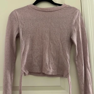 Glittrig ljuslila croppad tröja - Säljer en glittrig ljuslila croppad tröja med långa ärmar och snörning i midjan. Tröjan är ribbad och har en mjuk, stretchig känsla som sitter snyggt på kroppen. Den har även band som går att knyta runt midjan.