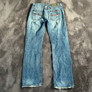 Nudie Jeans  - Säljer ett par klassiska blå jeans från Nudie Jeans med snygga slitningar. Jeansen har orange broderi på bakfickorna och råa, fransiga benslut. Tillverkade i bomull och har en autentisk vintagekänsla. Byxorna svänger ut där nere lite. Storlek 32/34