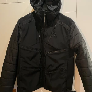 Svart jacka från C.P. Company - Säljer en svart pufferjacka från C.P. Company med huva och flera dragkedjefickor framtill. Jackan är tillverkad i återvunnen nylon och dun ärmar och rygg. Perfekt för kalla dagar och har en clean, modern look.