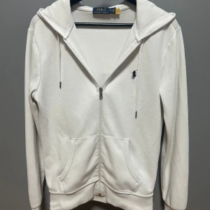 Vit hoodie från Polo Ralph Lauren XS - Vit zip hoodie från Polo Ralph Lauren i mjuk bomull. Klassisk design med dragkedja, huva med snörning och känguruficka framtill. Liten broderad logga på bröstet. Perfekt för en chill och stilren look. Endast en liten skråma på höger ärm som inte syns när man har tröjan på. 