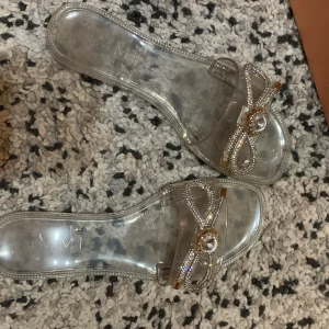 Glittriga sandaler från Simmi London - Säljer ett par transparenta sandaler från Simmi London i storlek 36. Skorna har en platt sula och är dekorerade med glittriga strassrosetter och en stor kristall framtill. Perfekta för dig som vill sticka ut med lite extra bling.