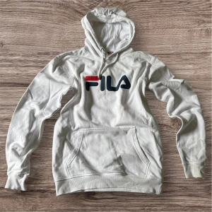 Vit hoodie från FILA med logga - Snygg vit hoodie från FILA med stor logga i rött och marinblått på bröstet. Klassisk känguruficka framtill, huva med snörning och mjukt bomullsmaterial. Perfekt för en chill och sportig look.