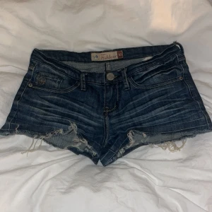 Mörkblå lågmidjade jeansshorts från Fishbone XS - Snygga mörkblå jeansshorts från Fishbone i storlek XS. Modellen är lågmidjad med slitna kanter och fransar för en cool, avslappnad look. Klassiska fickor fram och bak, samt peace-märke på myntfickan. Perfekta för sommardagar och festivaler.