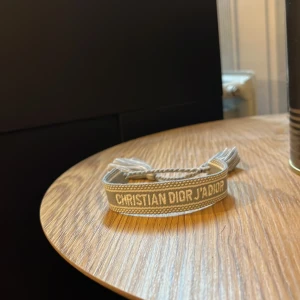 Grått armband från Christian Dior - Snyggt grått armband från Christian Dior med texten 'CHRISTIAN DIOR J'ADIOR' broderad i vitt. Armbandet är gjort i tyg med justerbar knytning och tofsar i ändarna. Perfekt accessoar för att lyfta din outfit med en lyxig touch. Äkta 