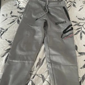 Grå mjukisbyxor från Hoodrich - Snygga grå mjukisbyxor från Hoodrich med svarta detaljer och tryck på ena benet. Byxorna har dragsko i midjan, fickor med dragkedja och en loose passform. Perfekta för en chill och sportig stil.