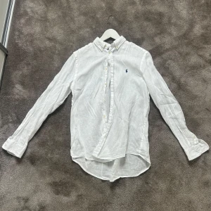 Vit linneskjorta från Ralph Lauren - Klassisk vit skjorta i linne från Ralph Lauren med button down-krage och broderad blå logga på bröstet. Långärmad modell med knappar framtill och rundad nederkant. Perfekt för en clean och stilren look. Skjortan är XL i barnstorlek vilket jag skulle säga är runt XS-S