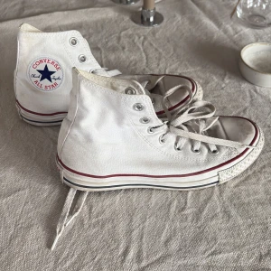 Vita Converse All Star high tops - Klassiska vita Converse All Star high tops med röd och blå rand runt sulan. Skorna har snörning, rund tå och är tillverkade i canvas. Perfekta för en avslappnad och cool stil. Ikonisk logga på sidan och slitstark gummisula.