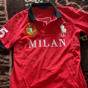 Röd pikétröja Polo Ralph Lauren Milan - Röd pikétröja från Polo Ralph Lauren med svart krage, stora broderade detaljer och texten MILAN framtill. Nummer 5 på ärm och rygg, samt klassisk polospelare-logga. Perfekt för dig som gillar sportig och stilren vibe.