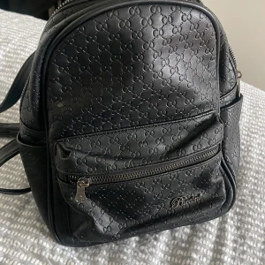 Svart ryggsäck från Puccini i skinn - Snygg svart ryggsäck från Puccini i skinnimitation med präglat mönster och silverfärgad dragkedja. Väskan har flera fack, både invändigt och utvändigt, och är perfekt för dig som vill ha stil och funktion i ett.