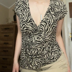 Zebramönstrad omlottopp från Indiska - Snygg omlottopp från Indiska i zebramönster med svart och beige. Djup v-ringning och lätt rynkade sidor ger en cool och trendig look. Tunn och mjuk, perfekt till jeans eller kjol. Kortärmad och figurnära passform.