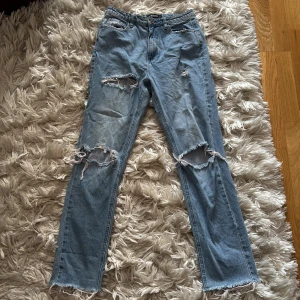 Blå ripped jeans från Missguided - Säljer ett par ljusblå jeans från Missguided med raka ben och slitna detaljer över knäna. Jeansen har fransade benslut och klassisk femficksdesign. Perfekta för en avslappnad och trendig look.