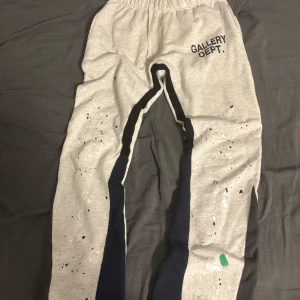 Grå mjukisbyxor från Gallery Dept. - Snygga grå mjukisbyxor från Gallery Dept. med svart logga på höften och coola färgstänk i vitt, svart och grönt. Byxorna har elastisk midja och svarta paneler längs benen. Perfekta för en avslappnad streetwear-look.