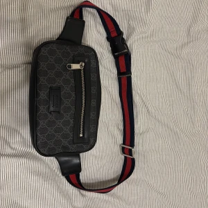 Svart axelväska monogrammönster  - Snygg svart axelväska frånmed klassiskt monogrammönster i grå toner. Väskan har en justerbar axelrem i rött och blått, dragkedja framtill och detaljer i skinn. Perfekt för dig som vill ha en stilren och lyxig accessoar.