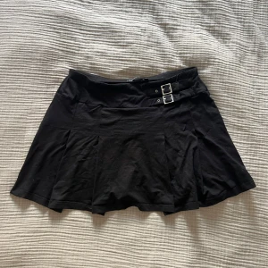 Svart minikjol med dubbla spännen från KappAhl - Svart kortkjol från KappAhl med två snygga silverspännen i midjan. Kjolen har en lätt utsvängd form och är tillverkad i ett mjukt tyg som känns bekvämt. Perfekt för dig som gillar en stilren och edgy look.