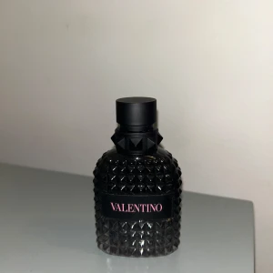 Valentino parfymflaska born in Roma  - Säljer en lyxig parfymflaska från Valentino i en svart, noppig glasflaska med elegant design och svart kork. Flaskan har en modern look med Valentino-loggan i rosa text på svart etikett. Perfekt för dig som gillar exklusiva dofter.50ml