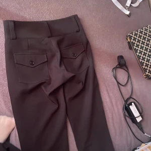 Svarta kostymbyxor från Gina Tricot - Säljer ett par svarta kostymbyxor från Gina Tricot med två knappar i midjan och fickor med lock och knapp bak. Byxorna är utsvängda. Helt nytt