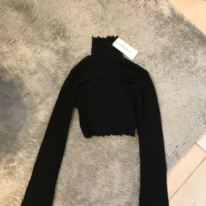 Svart croppad polotopp från Chiquelle - Säljer en svart croppad polotopp från Chiquelle. Toppen har långa ärmar och en hög krage med vågig kant. Perfekt att styla med höga jeans eller kjol för en trendig look.