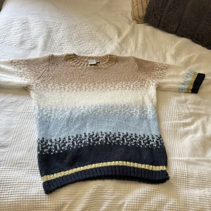Stickad tröja i beige, blå och mörkblå toner - Säljer en stickad tröja med rund hals och korta ärmar. Tröjan har ett snyggt mönster i beige, vitt, ljusblått och mörkblått med en guldig rand längst ner. Perfekt för kyliga dagar och riktigt mysig!