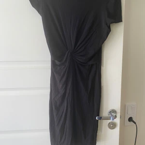 Svart klänning med knutdetalj i midjan - Säljer en svart klänning med kort ärm och snygg knutdetalj i midjan som ger en cool twist. Klänningen har en rak passform och är tillverkad i ett mjukt material som känns skönt mot huden. Perfekt för dig som gillar stilrena plagg med en liten detalj.