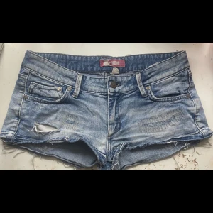 Ljusblå jeansshorts med slitningar från &NOW - Säljer ett par KORTA  ljusblå jeansshorts från &NOW med slitna detaljer och råa kanter. Shortsen har klassisk femficksdesign, väldigt låg midja och knappgylf. Perfekta för varma dagar och en avslappnad stil.  Midjemått: 40cm tvärs över. Skicka gärna prisförslag!!