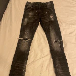 Svarta skinny jeans med slitningar från Supply & Demand - Säljer ett par svarta skinny jeans från Supply & Demand med slitningar och små dekorstenar framtill. Jeansen har klassisk femficksdesign, dragkedjegylf och är i storlek W30. Perfekta för dig som gillar en edgy stil.