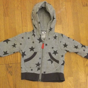 Åhléns grå hoodie, storlek 62-68 - Grå hoodie för barn från Åhléns med svarta stjärnor, huva och dragkedja med röd detalj. Mjukt bomullsmaterial och muddar vid ärmslut och midja.  Den har en röd detalj på dragkedjan och två fickor fram. För 4-6 månader Litet hål, se bild 4! Katter finns i hemmet!