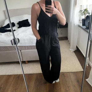 Flowig svart jumpsuit - Sval, tunn, flowig jumpsuit från H&M i strl 38. Liten öppning i ryggen och justerbara axelband, fickor på båda sidor.