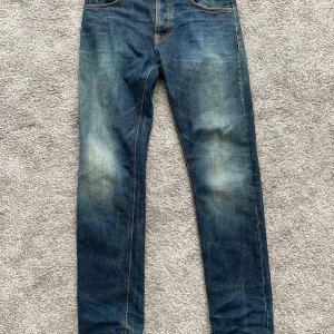 Jeans från Nudie Jeans - Säljer ett par klassiska blå jeans från Nudie Jeans med orangea sömmar och fem fickor. Jeansen har normal passform och raka ben, samt detaljerad logga på bakfickan och läderpatch i midjan. 30 waist 32 inseam