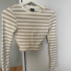 Beige och vit randig croppad tröja från Gina Tricot - Säljer en croppad långärmad tröja från Gina Tricot i beige och vitt randigt mönster. Tröjan har rund halsringning och är i ett mjukt, ribbat material. Perfekt att styla med höga jeans eller kjol.