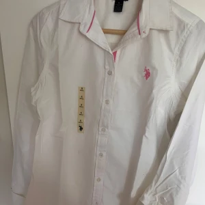 Vit skjorta från U.S. Polo - Klassisk vit skjorta från U.S. Polo Assn. med rosa broderad logga på bröstet och rosa detaljer vid kragen. 