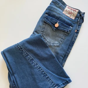 Blå jeansbyxor från True Religion - Säljer ett par klassiska blå jeans från True Religion med snygga kontrastsömmar och dekorativa fickor bak. Modellen heter Joey och har midwaist.