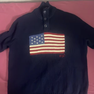 Mörkblå stickad tröja med amerikansk flagga från Polo Ralph Lauren - Mörkblå stickad tröja från Polo Ralph Lauren med stor amerikansk flagga framtill och knappslå vid halsen. Tröjan har långa ärmar och ribbade muddar. Klassisk och stilren design med RL-broderi under flaggan.