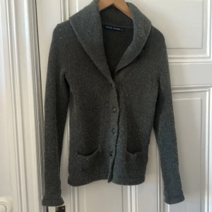 Grå kofta från Ralph Lauren - Mysig grå kofta från Ralph Lauren med knappar framtill och två fickor. Koftan har en bred krage och är tillverkad i en mjuk blandning av bomull och kashmir. Perfekt för lager-på-lager under kyliga dagar.