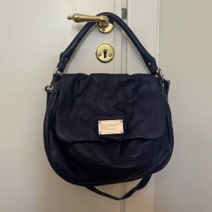 Mörkblå axelväska från Marc by Marc Jacobs - Säljer en snygg mörkblå axelväska från Marc by Marc Jacobs med silverfärgade metalldetaljer och logoplatta framtill. Väskan har ett stort huvudfack och ett coolt svartvitt innerfoder med text. Perfekt för dig som vill ha en stilren och rymlig väska.
