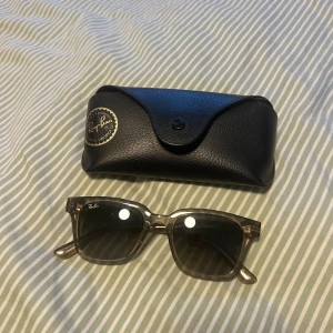 Ray-Ban solglasögon med svart fodral - Snygga Ray-Ban solglasögon med transparent grå båge och mörka glas. Klassisk fyrkantig form och diskret Ray-Ban-logga på linsen. Kommer med svart fodral och putsduk. Perfekt accessoar för soliga dagar.