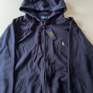Mörkblå  hoodie med dragkedja från Polo Ralph Lauren - Säljer en svart hoodie från Polo Ralph Lauren med dragkedja framtill och huva med snörning. Klassisk logga broderad på bröstet och två fickor framtill. Perfekt för en avslappnad stil. (Helt ny,Oanvänd)