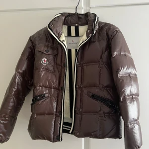 Brun dunjacka från Moncler - Säljer en brun, glansig dunjacka från Moncler med svartvita detaljer vid kragen. Jackan har dragkedja, två snedställda fickor med dragkedja och en bröstficka med knapp. Klassisk Moncler-logga på bröstet och quiltad design. Den är liten sliten då den är ganska gammal men går att använda. Har tyvärr inget kvitto då det är min mammas gamla. Tveka inte att höra av dig vid frågor!