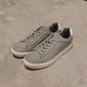 Grå sneakers med vit sula - Snygga grå sneakers med vit sula och vit detalj på hälen. Skorna har snörning och är tillverkade i mocka-liknande material. Perfekta för en stilren och enkel look.