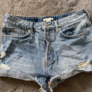 Blå jeansshorts - Snygga blå jeansshorts med slitna detaljer och fransig kant. Klassisk femficksmodell med knappar i gylfen och låg midja. Perfekta för sommaren och ger en avslappnad look.