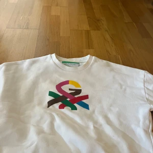 Vit sweatshirt från United Colors of Benetton - Vit sweatshirt med färgglad Benetton-logga på bröstet. Tröjan har rund halsringning och långa ärmar. Perfekt för en avslappnad och stilren look.