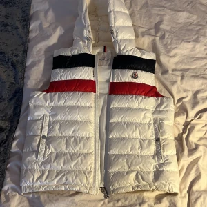Vit dunväst från Moncler med huva - Snygg vit dunväst från Moncler med huva och dragkedja. Västen har svarta och röda detaljer upptill samt Moncler-logga på bröstet. Två fickor framtill med tryckknappar. Perfekt för lager-på-lager under kyliga dagar. Dock en liten skada som knappt syns med ett vitt klister märke, priset kan diskuteras.
