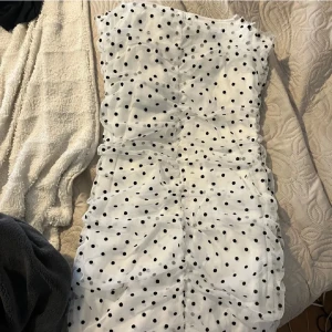 Vit prickig axelbandslös klänning - Säljer en vit axelbandslös klänning med svarta prickar och rynkade detaljer längs hela klänningen. Klänningen har en tight passform och är kort i modellen.