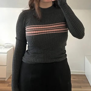 Mörkgrå ribbad tröja med rosa ränder - Säljer en mörkgrå ribbad tröja med rosa horisontella ränder över bröstet. Tröjan har lång ärm, hög hals och figurnära passform. Aldrig använd.
