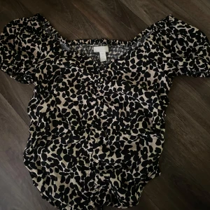 Leopardmönstrad croppad blus från H&M - Säljer en croppad blus från H&M med leopardmönster i svart och beige. Blusen har korta puffärmar, smockad rygg och knytning framtill. Perfekt för dig som gillar trendiga och djurmönstrade plagg. Den är aldrig använd då den inte passar mig🐆🤍