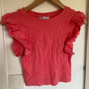 Rosa volangtopp från Zara - Supersöt rosa topp från Zara med volangdetaljer längs axlarna. Toppen är ribbad och har en rund halsringning. Perfekt för dig som vill ha en färgglad och trendig look. Storlek S men passar som en XS. Perfekt för neon fest!