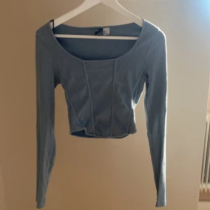 Ljusblå croppad långärmad topp - Säljer en ljusblå croppad topp med långa ärmar och fyrkantig urringning. Toppen är ribbad och har markerade sömmar framtill för en snygg detalj. Perfekt att styla med jeans eller kjol.