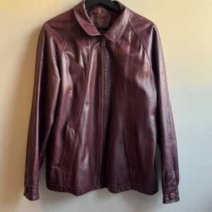 Vinröd skinnjacka  - Säljer en vinröd skinnjacka med klassisk krage och dragkedja framtill. Jackan har två snedställda fickor och knappdetalj vid ärmslutet.