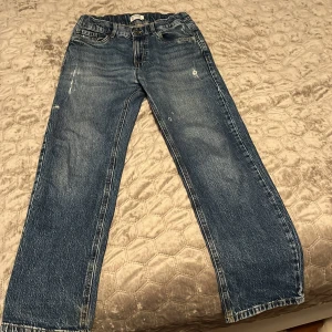 Blå raka jeans med slitningar från Lindex  - Klassiska blå jeans med raka ben och lätt slitna detaljer framtill. Jeansen har fem fickor, normal passform och stängs med dragkedja och knapp. Perfekta för en avslappnad stil.